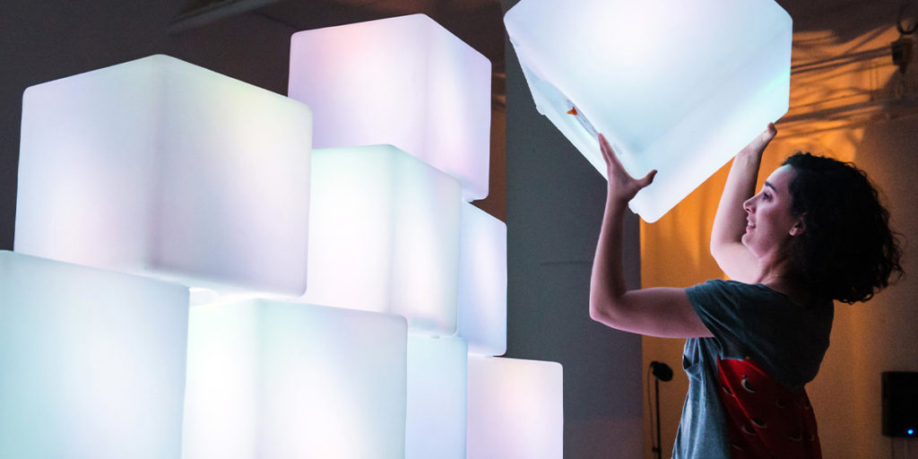 A woman with curly hair wearing a colorful dress lifts a glowing cube, ready to stack it on the top of a wall of similar glowing cubes which make up Sound Sculpture.