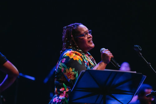 PaviElle French sings while holding a microphone, wearing a colorful top and glasses. A music stand is in the foreground.