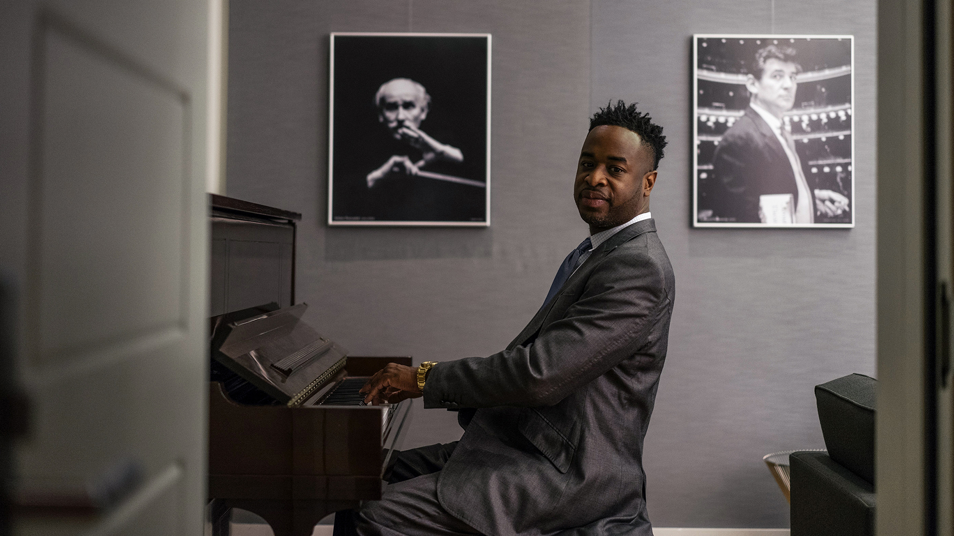 Damien Sneed, composer and pianist, sits on a piano bench with his hands over the keys. His face is turned towards the camera, and he is wearing a dark grey suit. The wall behind him is grey and two photographs hang on the wall.