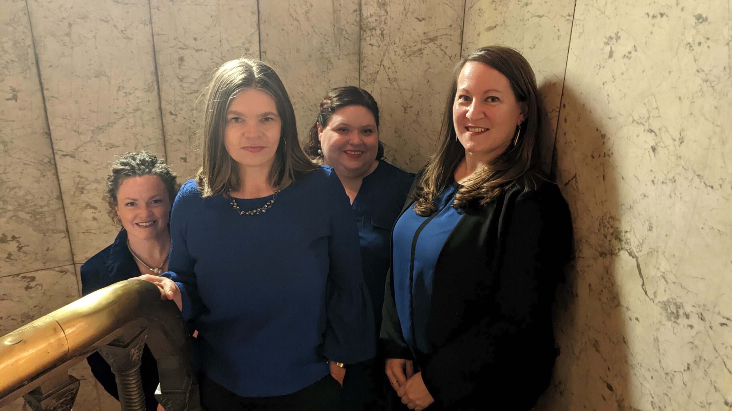 The four members of Lumina Women's ensemble stand together in a stairway, all smiling at the camera. Two women are in the foreground and two staggered behind them on a lower stair. All four women are wearing blue.