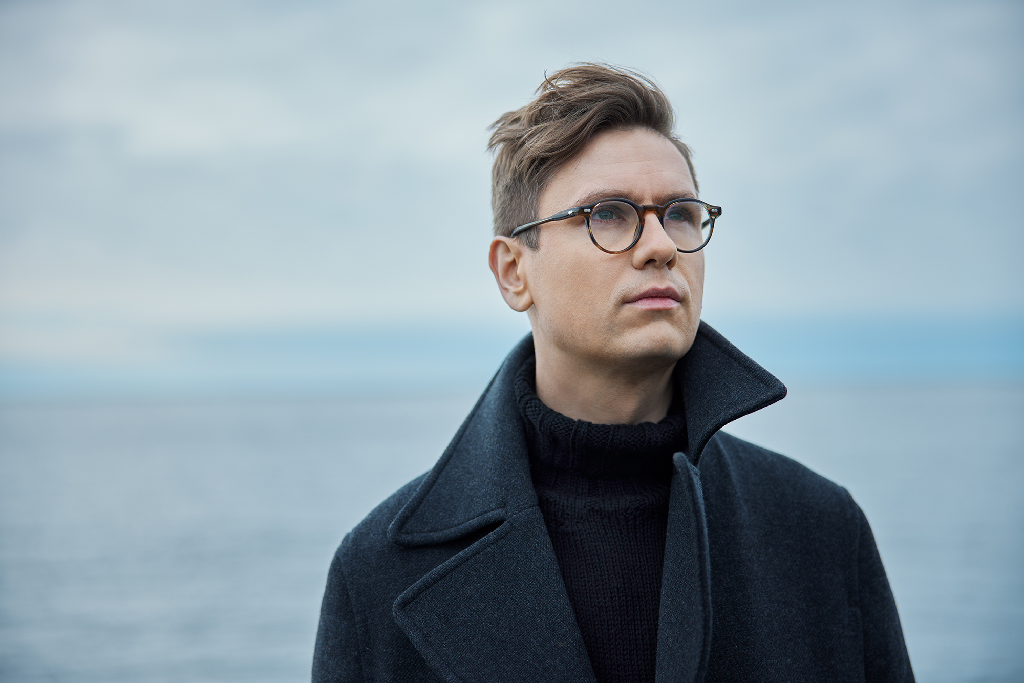 Pianist Vikingur Olafsson stands in front of the ocean, gazing upward and slightly to the right. He is wearing glasses and a dark shirt and coat.